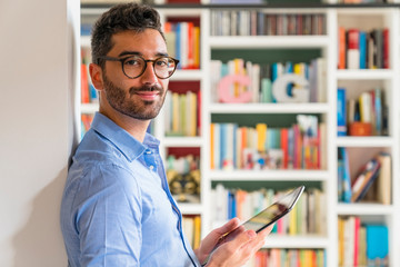 Portrait of smiling young man standing in front of bookshelves at home with digital tablet