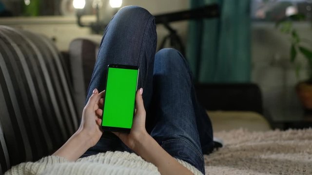 young girl is resting at home on the couch and holding a smartphone with a green screen. green mockup to insert apps or images. Internet addiction. Gesture flipping photos with your fingers