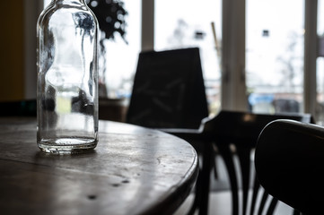 Empty, transparent bottle of drinking water stands on a round wooden table, illuminated by daylight, on a blurry background of large windows, in a cafe.