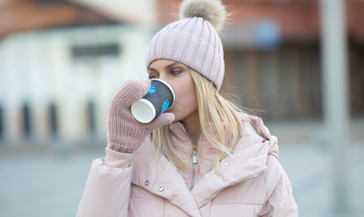 Cute young Caucasian teenage woman in beige  hat with pompon and pink mittens holding steaming cup of hot tea or coffee, outdoor in sunny winter day.