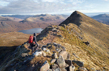 A hiker walking over the narrow ridge that leads to Sgurr an Tuill Bhain with Lochan Fada in the distance in the Scottish Highlands.