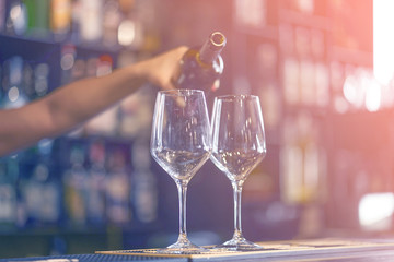 Hand a barman girl pours white wine into a glass from a bottle. Shelves with bottles of alcohol in the background. Focus on the glass. Toning