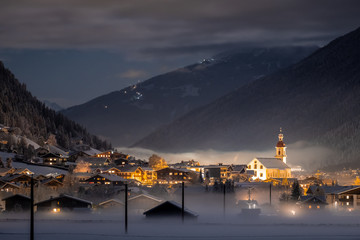 Winter night landscape overlooking the Austrian Tyrolean city of Neustift and the Pfarre church against the backdrop of mountains and clouds. Frosty night with fog in the valley