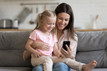 Child daughter sit on mom lap on sofa using phone
