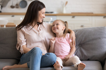 Happy young adult mum and little daughter talking on sofa