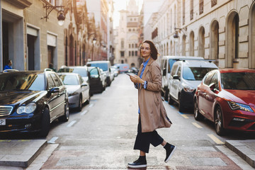 Fototapeta premium Stylish tourist girl walking on the old european streets with offline map on smartphone, Smiling female student in stylish clothing messaging on smartphone while walking