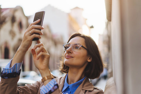 From below of modern woman in trench coat and glasses smartphone looking around while standing on city street, social media concept, Woman taking pictures on smartphone while walking on the street