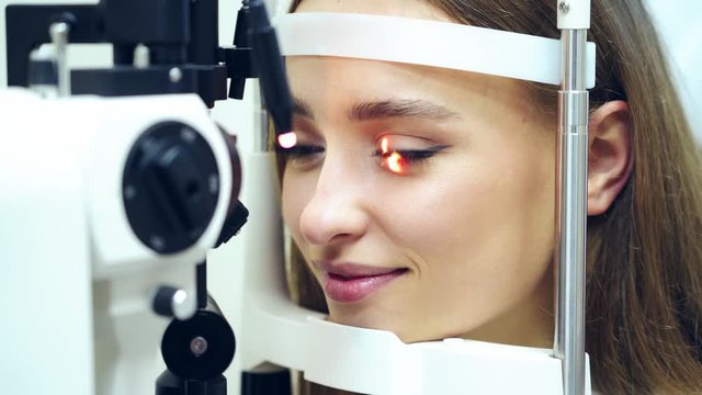 Face of a young female at an ophthalmologist office. Smiling woman holds eyes open during an optical exam. Eye check up by modern microscope. Medical device laser shine inside the eye.
