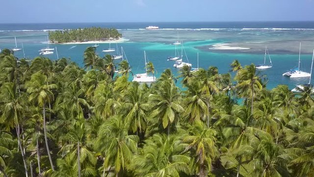 A nice drone shot flying over green palm trees to reveal many sailboats anchoring in a blue lagoon between two tropical islands on a sunny day in San Blas, Panama.
