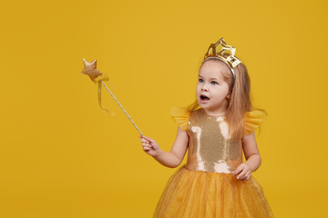 Joyful little girl with long hair in a tulle golden dress and princess crown holding a magic wand ...