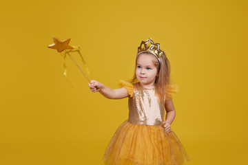 Joyful little girl with long hair in a tulle golden dress and princess crown holding a magic wand ...