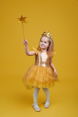 Joyful little girl with long hair in a tulle golden dress and princess crown holding a magic wand  on yellow background. Celebrating a colorful carnival for kids, expressing positive birthday