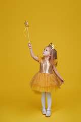 Joyful little girl with long hair in a tulle golden dress and princess crown holding a magic wand ...