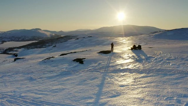 Drone flies low over a standalone person and snowmobile up towards golden Sun just before the sunset time. Winter mountains at Atoklinten, Joesjo, Lappland, Northern Sweden