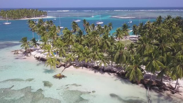A beautiful drone shot orbiting around a tropical island with white sand beaches, covered by green palm trees in San Blas, Panama. Lots of sailboats anchoring in a blue lagoon between the islands.
