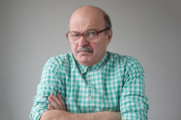 Angry senior man in glasses crossing arms looking annoyed at camera. He does not trust the news he...