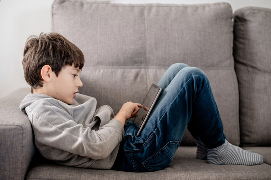A boy concentrated playing with a tablet layed down in a sofa