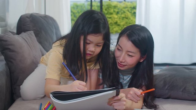 Asian middle-aged woman teach her daughter do homework and drawing at home. Lifestyle mother and kid happy fun spend time together in living room in modern home in the evening concept.