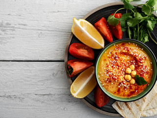 Bowl with traditional homemade hummus with olive oil and fresh vegetables. Slices of lemon and pita nearby on a plate. Useful natural snacks. Background of wooden white boards. Top view. Copy space