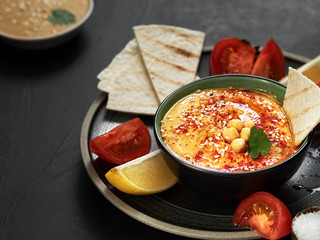Bowl with traditional hummus with olive oil and fresh vegetables and a bowl with tahini. Slices of lemon and pita nearby on a plate. Useful natural snacks. Black stone background. Close-up.