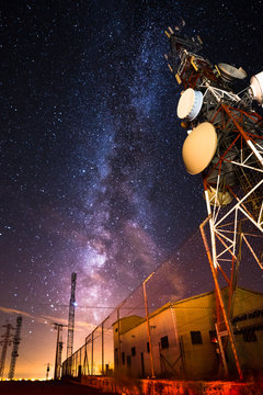 From below wonderful view of media antenna and silhouettes of electricity high voltage power towers against night city on horizon and gorgeous Milky Way in clear sky