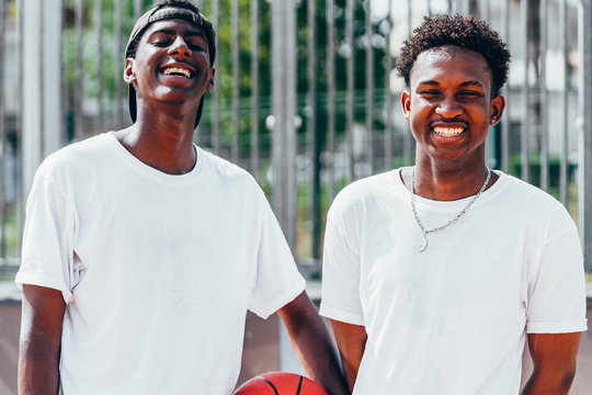 happy African American guys holding orange ball and looking at camera in bright day near metal fence