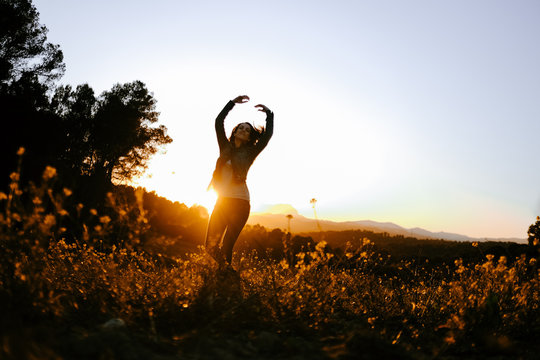 Relaxed young female in black jacket with outstretched arms enjoying nature and freedom while walking on beautiful meadow on sunset behind trees