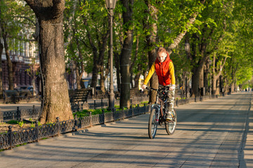 A boy of 8 years old in an orange vest rides a bicycle in a city park on a sunny spring morning.