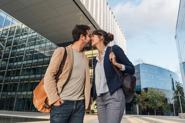 From below happy couple coworkers kissing each other while standing outside modern building on city street after work