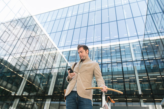 From below of pensive adult male in casual clothes making phone call while standing with bike against contemporary high rise business center with glass walls in downtown