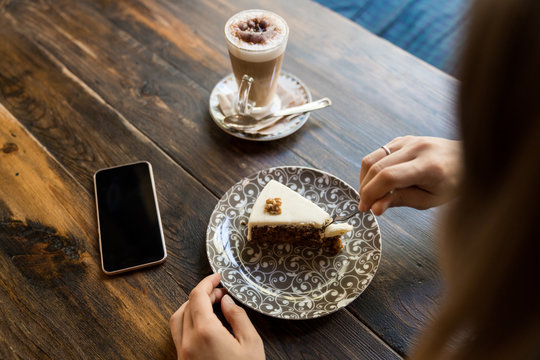 Female holding fork and sitting at table with blank screen smartphone next to coffee and cake on plate with ornament