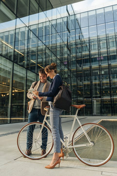 Cheerful man and woman with bicycle smiling and looking at a tablet while communicating outside office building on modern city street