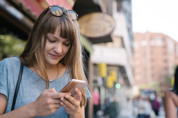 Blonde young cheerful female with sunglasses in blue T-shirt standing on street of big city and typing message on smartphone