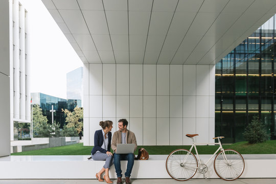 Delighted businesspeople smiling and browsing laptop together while sitting outside modern building near bicycle on city street