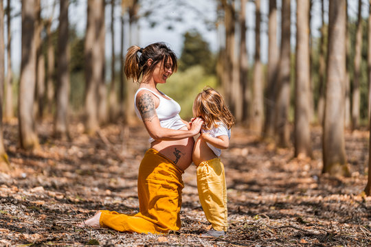 Side view of cheerful barefooted pregnant woman and little girl in similar clothes holding hands and touching bellies to each other while having fun in autumn forest glade during sunny day