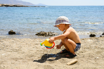a cute little three year old boy in a sun hat squats and plays on the seashore against the backdrop of the seascape