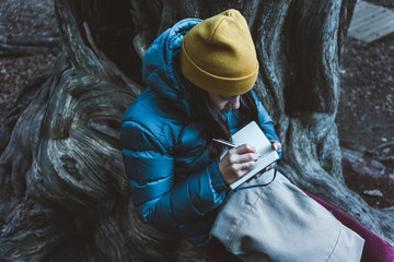 From above of teenage girl in warm active wear with backpack sitting on massive roots of old huge tree and taking notes in small notebook during exciting trekking in autumn forest