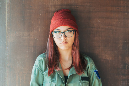 Thoughtful stylish woman in warm hat and glasses in dark green shirt nearby brown wall looking along