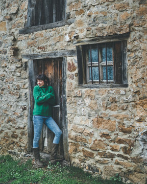 Casual female in green sweater and jeans standing with crossed arms next to door of old stone country house and feeling cold on gloomy damp autumn day