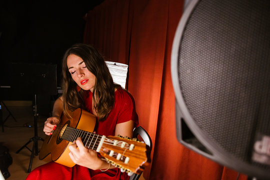 Charming artistic woman with closed eyes in red dress performing song playing on guitar in stage with warm light in Spain