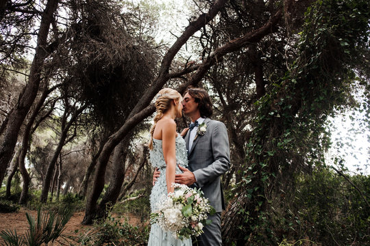 Young newly wedded couple in wedding wear standing and hugging on pathway among beautiful green forest with big trees