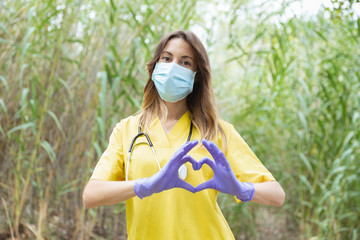 Mujer enfermera joven vestida con uniforme amarillo, guantes y mascarilla, forma un corazón con...