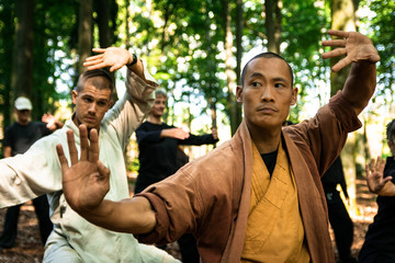 Ethnic trainer in uniform gesticulating and looking away while showing exercise to group of students during martial arts workout in forest