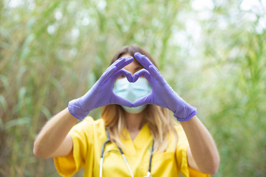 Mujer enfermera joven vestida con uniforme amarillo, guantes y mascarilla, forma un coraz&oacute;n con sus manos en entorno de naturaleza