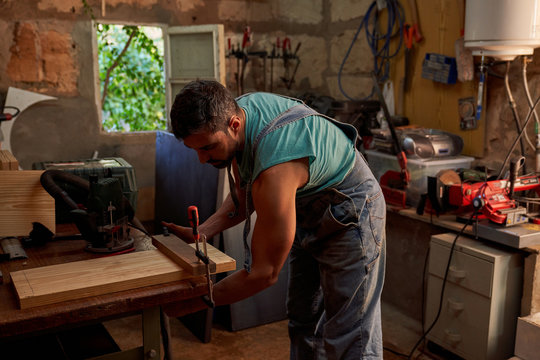 Side view of skilled middle aged handyman in overalls using clamp while working with wooden plank in workshop - Powered by Adobe