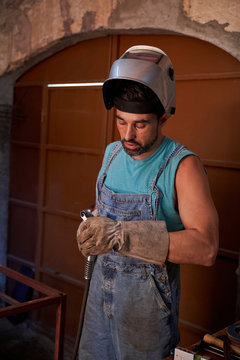 Professional man with welding mask on head wearing jeans overalls and protective gloves standing on workplace and preparing instrument for soldering