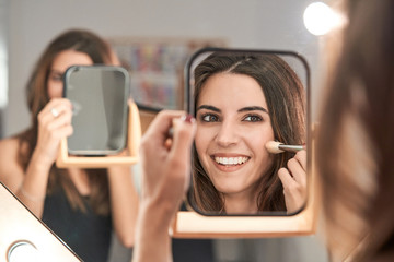 Long haired woman making up on face drawing blush on cheeks with brush looking at mirror in hand in light stylish salon
