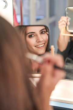 Long haired woman making up on face drawing blush on cheeks with brush looking at mirror in hand in light stylish salon