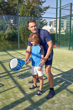 Adult man showing boy how to hold paddle and hit ball during tennis training on sunny day on court