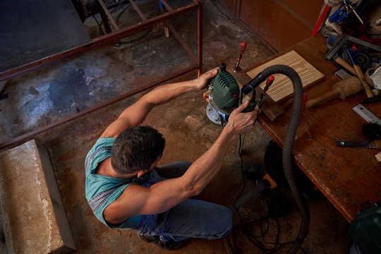 From above of male worker using milling machine while processing wooden plank on workbench in workshop